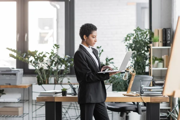 現代のオフィスのワークデスクに立ちながらノートパソコンを使う若いアフリカ系アメリカ人のビジネスマン — ストック写真