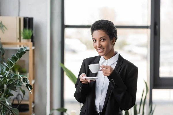 快乐的非洲裔美国女商人一边拿着茶托和咖啡杯 一边对着办公室里的摄像机微笑 — 图库照片