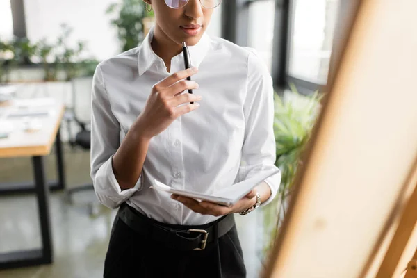 Stock image cropped view of african american businesswoman in white blouse standing with pen and notebook near blurred note board in office