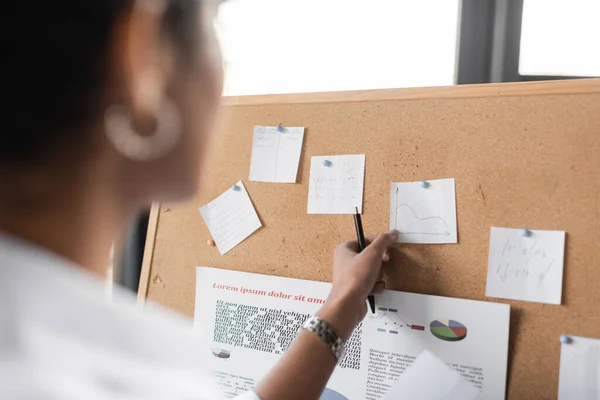 アフリカ系アメリカ人のビジネス女性がオフィスのコルクボード上のインフォグラフィックで紙のノートに触れる — ストック写真
