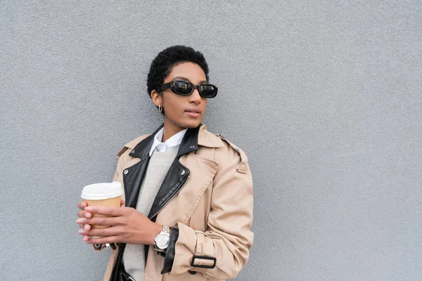 stock image african american businesswoman in beige trench coat and sunglasses holding paper cup and looking away near grey wall in city