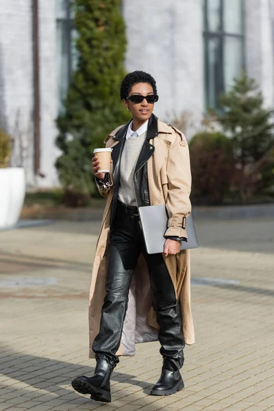 Stock image african american businesswoman in beige trench coat and sunglasses walking with disposable cup and laptop on blurred city street