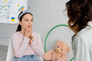 Preteen kid sticking out tongue during lesson with speech therapist in consulting room  clipart