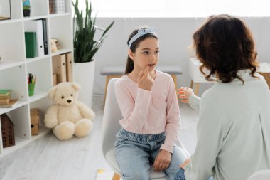 Preteen girl talking and pointing at mouth near blurred speech therapist in consulting room  clipart