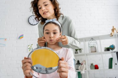 Speech therapist touching cheeks of pupil with blurred mirror in consulting room  clipart