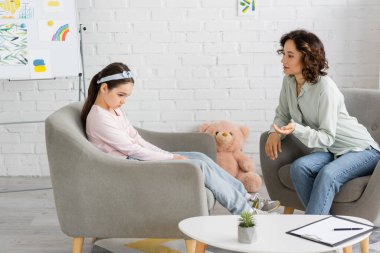 Psychologist talking to sad preteen girl on armchair in consulting room  clipart