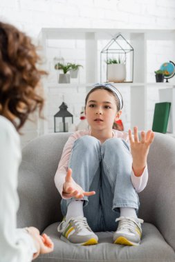 Preteen girl talking to blurred psychologist while sitting on armchair in consulting room  clipart