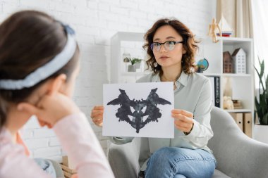 Gözlüklü psikolog Rorschach testini muayene odasında bulanık bir çocuğun yanında tutuyor. 