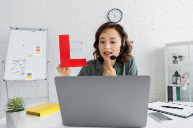 Speech therapist in headset holding letter and pointing at mouth during online lesson on laptop in consulting room  clipart