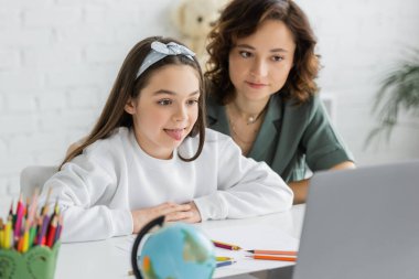 Online konuşma terapisi sırasında annemin yanında dilini dışarı çıkaran küçük kız. 
