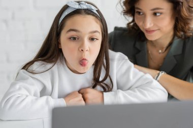 Laptopta konuşma terapisi dersi sırasında dilini dışarı çıkaran küçük bir kız evde bulanık bir annenin yanında. 