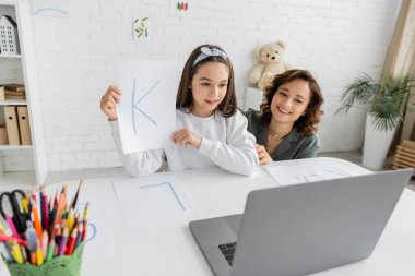 Evde konuşma terapisi videosu dersi alan, kızının yanındaki dizüstü bilgisayara bakan çok mutlu bir ebeveyn. 