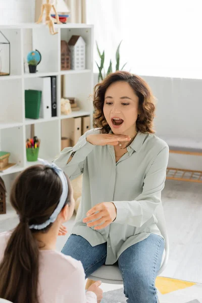 Speech therapist opening mouth and gesturing near blurred pupil in consulting room