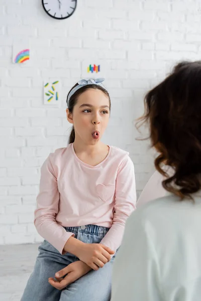 Preteen girl sticking out tongue near speech therapist in consulting room