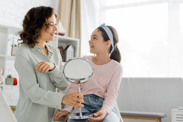 Smiling speech therapist holding mirror and looking at pupil in consulting room