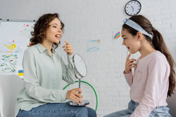 Speech therapist holding mirror and pointing at mouth near pupil in consulting room