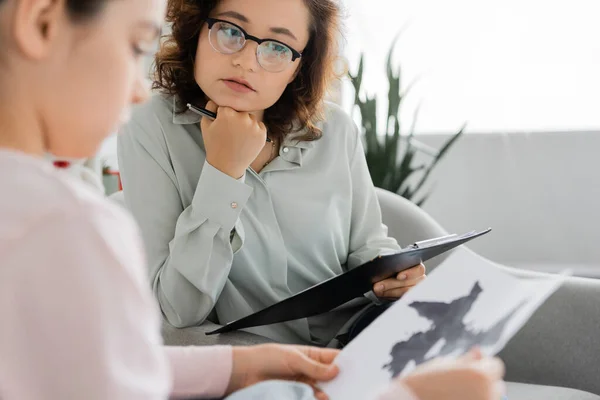 stock image Psychologist holding clipboard near blurred child with Rorschach test in consulting room 