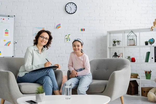 Cheerful Psychologist Writing Clipboard Looking Camera Preteen Girl Consulting Room — Stock Photo, Image