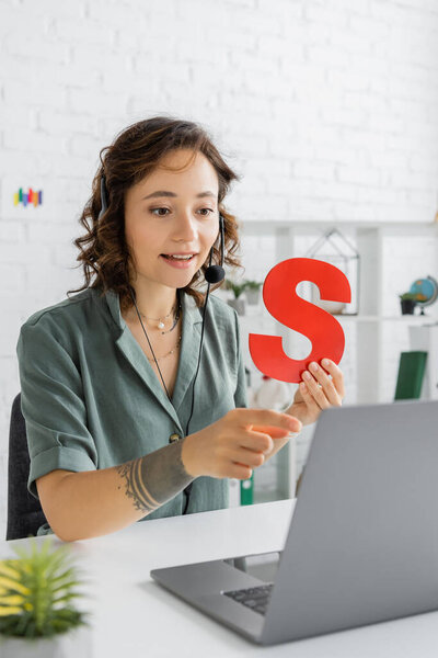 Улыбающийся логопед в наушниках, держащий буквы и указывающий на ноутбук во время видеозвонка в консультационной комнате 