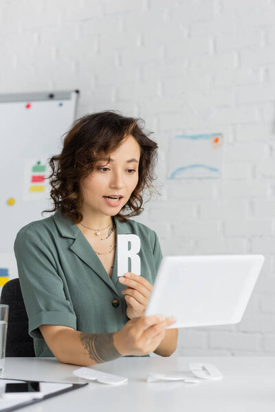 Татуированный логопед держит букву "б" и разговаривает во время видеозвонка на цифровом планшете в консультационном кабинете 