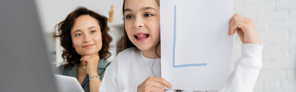 Ребенок торчит язык и держит бумагу с письмом во время логопедии онлайн рядом с матерью дома, баннер 
