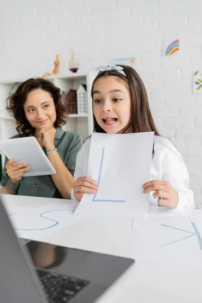 stock image Girl holding paper with letter during speech therapy video lesson near smiling mom with digital tablet at home 