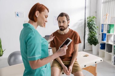 redhead physiotherapist looking at digital tablet near sad bearded man sitting on massage table in consulting room clipart