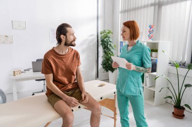 redhead rehabilitation specialist with digital tablet talking to bearded man sitting on massage table in consulting room clipart