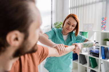 positive physiotherapist examining injured arm of blurred man in consulting room clipart