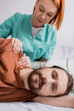 Kızıl saçlı manuel terapist rehabilitasyon merkezinde sakallı adamın acı dolu omzuna masaj yapıyor.