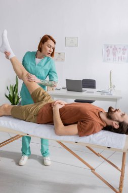 Fizyoterapist, hasta sakallı bir adamın bacağını geriyor. Masaj masasında yatıyor.
