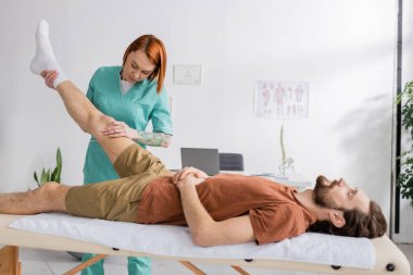 Kızıl saçlı manüel terapist rehabilitasyon merkezinde sakallı adamın acılı bacağına masaj yapıyor.