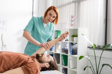 Rehabilitasyon merkezinde yaralı bir hastanın koluyla çalışırken kameraya bakan kızıl saçlı fizyoterapist.