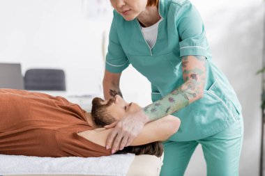 Dövmeli fizyoterapist rehabilitasyon merkezinde tedavi sırasında sakallı adamın acı dolu koluna masaj yapıyor.