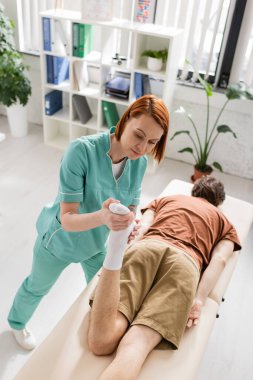 Klinikteki masaj masasında yatan hastanın ayakları üzerinde iyileşme manipülasyonu yapan kızıl saçlı kayropraktörün yüksek açılı görüntüsü.