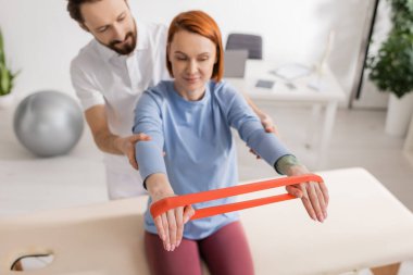 Klinikteki masaj masasında direniş bandıyla çalışan gülümseyen kadını destekleyen fizyoterapist.
