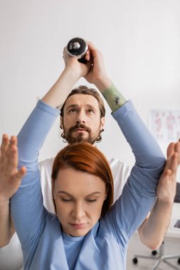 Sakallı rehabilitasyon uzmanı danışma odasında dambıllarla egzersiz yapan bir kadına yardım ediyor.