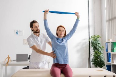 Sakallı rehabilitasyon doktoru danışma odasında masaj masasında oturan kadına yardım ediyor ve direniş bandıyla kollarını eğitiyor.