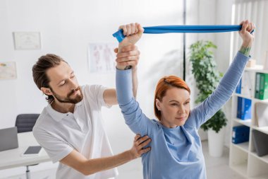 Rehabilitolog, danışma odasında direniş bandı eğitimi alan kızıl saçlı bir kadına yardım ediyor.