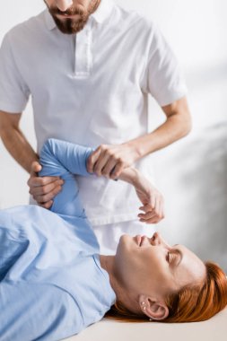 Sakallı fizyoterapist klinikteki rehabilitasyon sırasında kadının acı dolu kolunu esnetiyor.