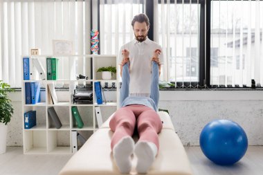 Sakallı fizyoterapist modern rehabilitasyon merkezinde masaj masasında uzanan kadının acı kollarını esnetiyor.
