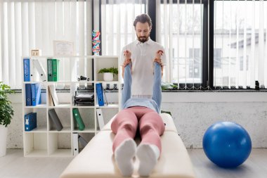 bearded physiotherapist stretching painful arms of woman lying on massage table in modern rehabilitation center clipart