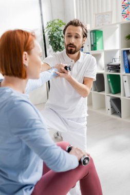 Sakallı fizyoterapist, tedavi kliniğinde, kızıl saçlı kadının halterlerle çalışmasına yardım ediyor.