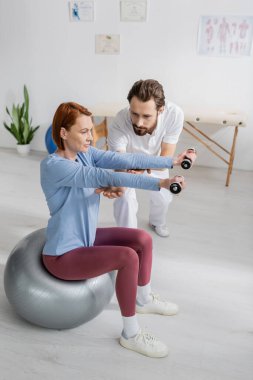 rehabilitologist helping redhead woman working out with dumbbells while sitting on fitness ball in rehab center clipart