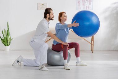 Sakallı fizyoterapist, klinikte rehabilitasyon sırasında kadının fitball çalışmasına yardım ediyor.