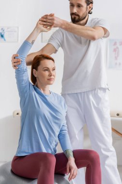 physiotherapist supporting arm of redhead woman training on fitness ball in recovery clinic clipart