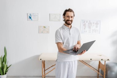 bearded rehabilitation specialist standing with laptop and looking at camera in consulting room clipart