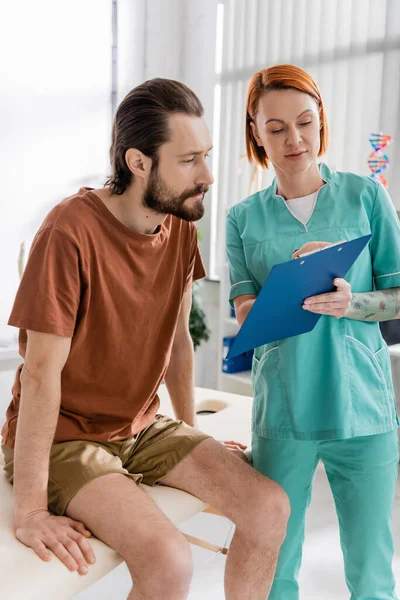 Physiotherapist Showing Prescription Clipboard Bearded Man Sitting Massage Table Consulting — Stock Photo, Image