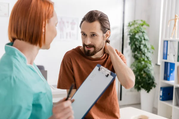 Bearded Man Touching Painful Neck Blurred Physiotherapist Clipboard Consulting Room — Stock Photo, Image