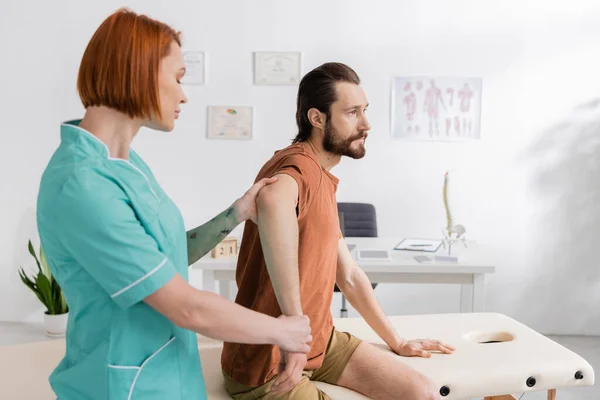 診察室での診察中に負傷者の腕を屈曲させている赤毛の理学療法士の側面図 — ストック写真